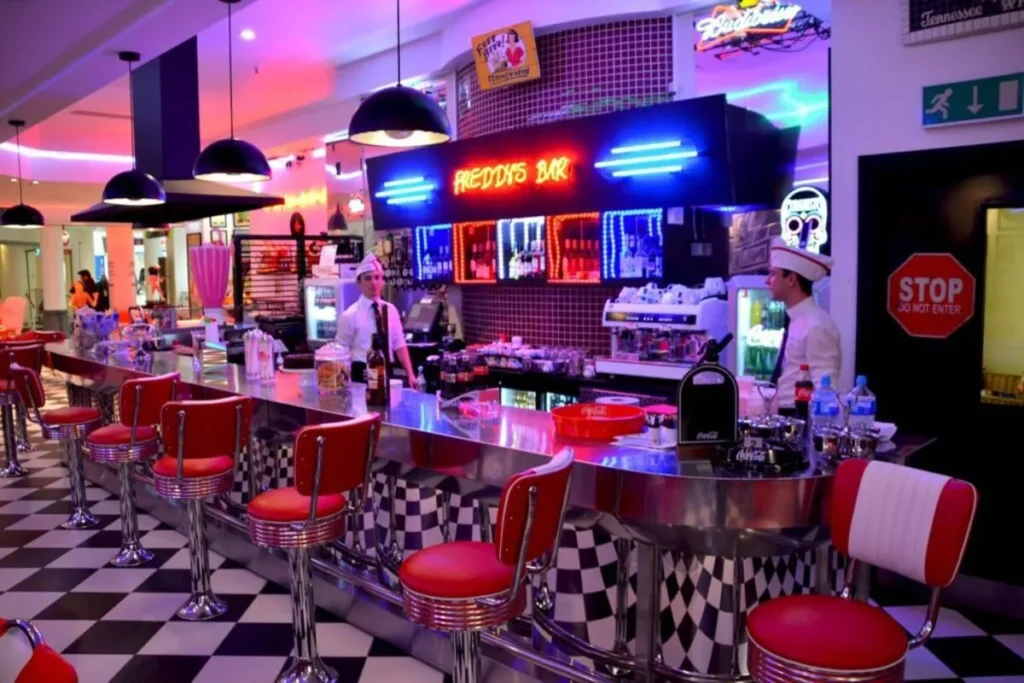 Interior de uma lanchonete retrô no estilo de um diner americano, com um balcão de metal, bancos altos vermelhos e piso quadriculado. A imagem retrata o ambiente de uma das muitas lanchonetes 24 horas que oferecem o estilo dos anos 50.
