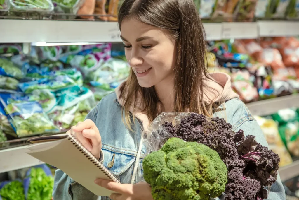 Mulher em supermercado, segurando couve-flor e couve roxa, enquanto verifica a sua lista de compras anotada em um caderno.
