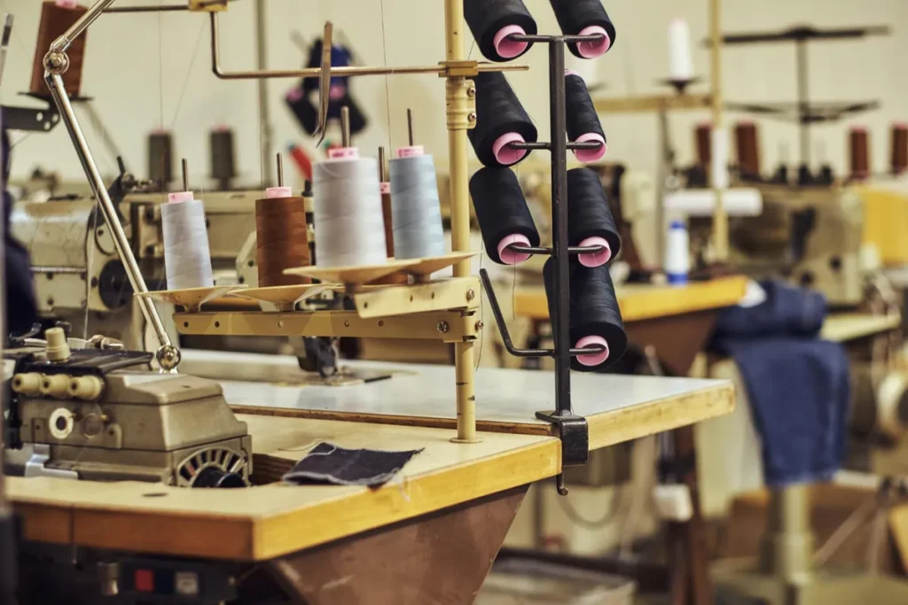 Máquina de costura industrial com vários carretéis de linha em cores diferentes, como preto, cinza e marrom, em uma oficina de confecção.