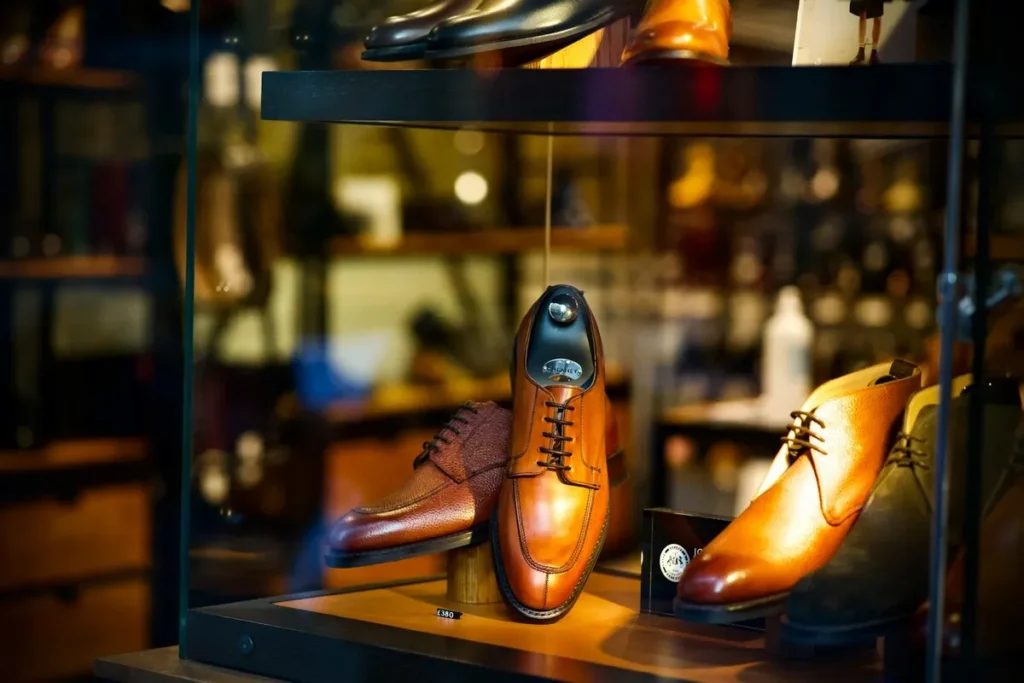 Sapatos sociais masculinos de couro (Oxford e Derby) em tons de caramelo e marrom em vitrine de loja de calçados de luxo com iluminação quente.