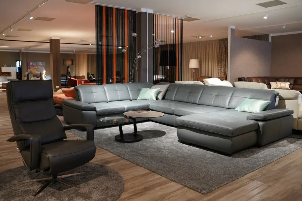 Grande sofá modular de canto em couro cinza-escuro com almofadas de destaque e poltrona reclinável preta em um showroom de móveis contemporâneos.