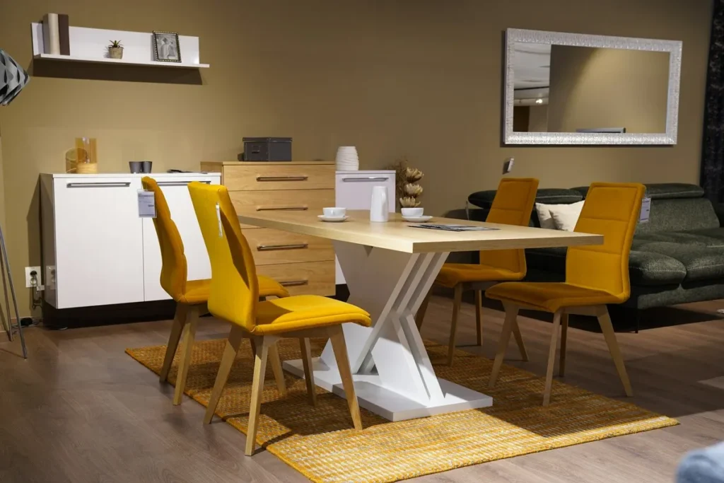 Conjunto de sala de jantar moderno com mesa de madeira clara e base branca em X, quatro cadeiras estofadas em veludo amarelo mostarda e tapete de sisal amarelo em um showroom de loja de móveis.