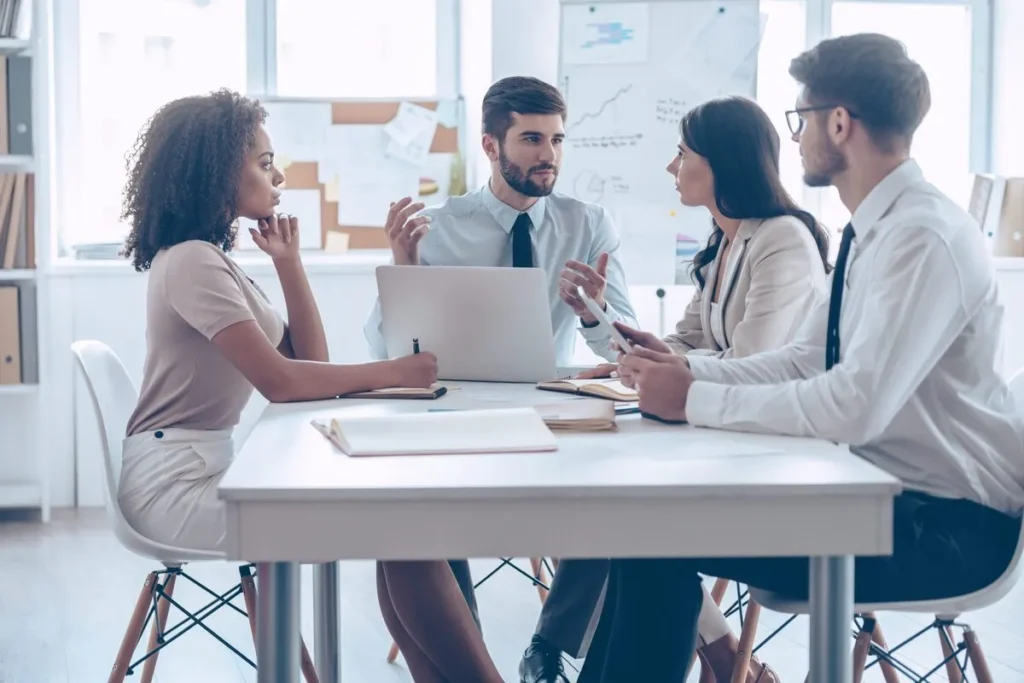 Quatro colegas de trabalho em uma reunião de equipe, discutindo e planejando novas metas. A imagem representa o trabalho em equipe e o planejamento estratégico em um ambiente de escritório.