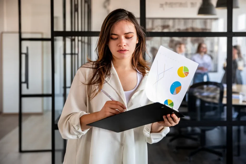 Uma mulher com uma camisa branca, em pé em um escritório moderno, segura uma prancheta e gráficos de pizza, analisando os resultados de uma pesquisa de clima organizacional.