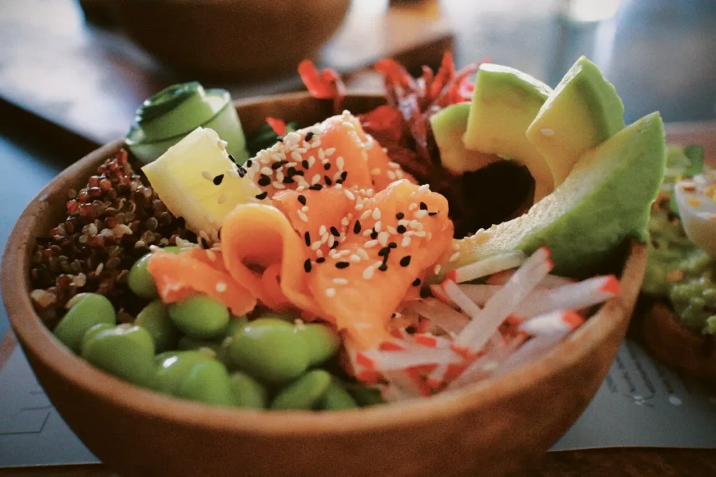 Close-up de Poke Bowl em tigela de madeira rústica com salmão em fatias, abacate, edamame, quinoa e pepino em rodelas, coberto com gergelim em foto com filtro vintage.