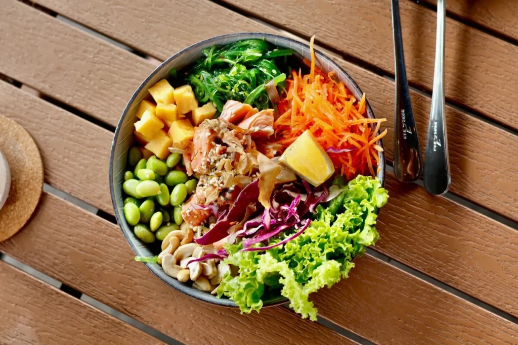 Vista de cima de um Poke Bowl colorido com salmão desfiado, manga em cubos, edamame, cenoura ralada, algas marinhas e castanhas de caju, servido sobre mesa de madeira.