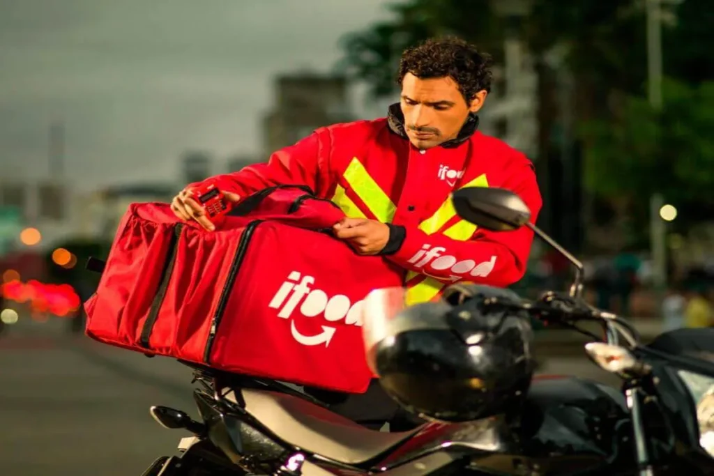 Entregador de aplicativo iFood vestindo jaqueta vermelha e mochila térmica vermelha com logo, colocando ou retirando pedido em cima de motocicleta na rua.