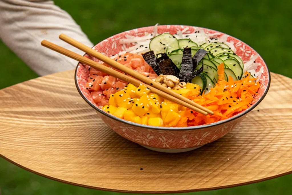 Close-up de Poke Bowl com salmão em cubos, manga, cenoura ralada, pepino, algas e hashi, sendo servida em tigela decorada sobre bandeja de madeira, com fundo de grama verde.