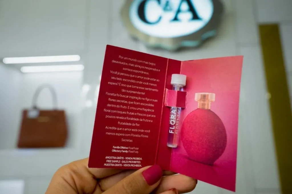 Uma pessoa segurando uma amostra grátis de perfume da linha Floratta. A imagem demonstra uma estratégia de marketing de sampling, ou amostragem, onde as empresas distribuem pequenas quantidades de produtos para potenciais clientes experimentarem.