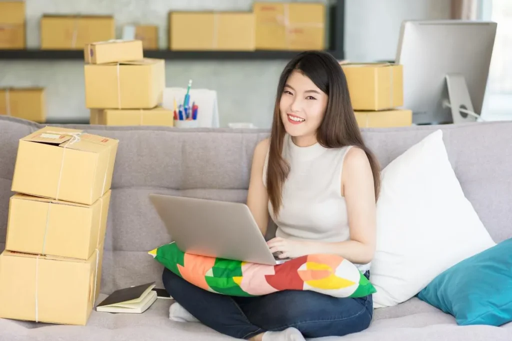 Uma jovem empreendedora de e-commerce sorrindo para a câmera, com caixas de encomendas ao seu redor. Ela usa um laptop para gerenciar as vendas e implementar estratégias de upsell e cross-sell.