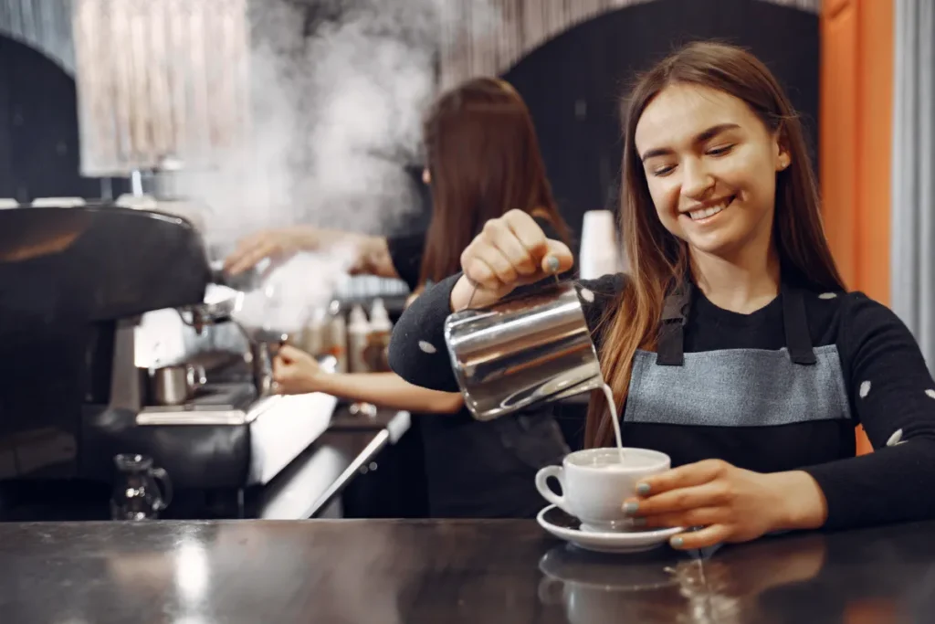 Barista (mulher sorridente) adicionando leite vaporizado para criar Latte Art em uma xícara de café. A cena mostra a preparação em uma cafeteria gourmet com máquina de espresso profissional ao fundo.