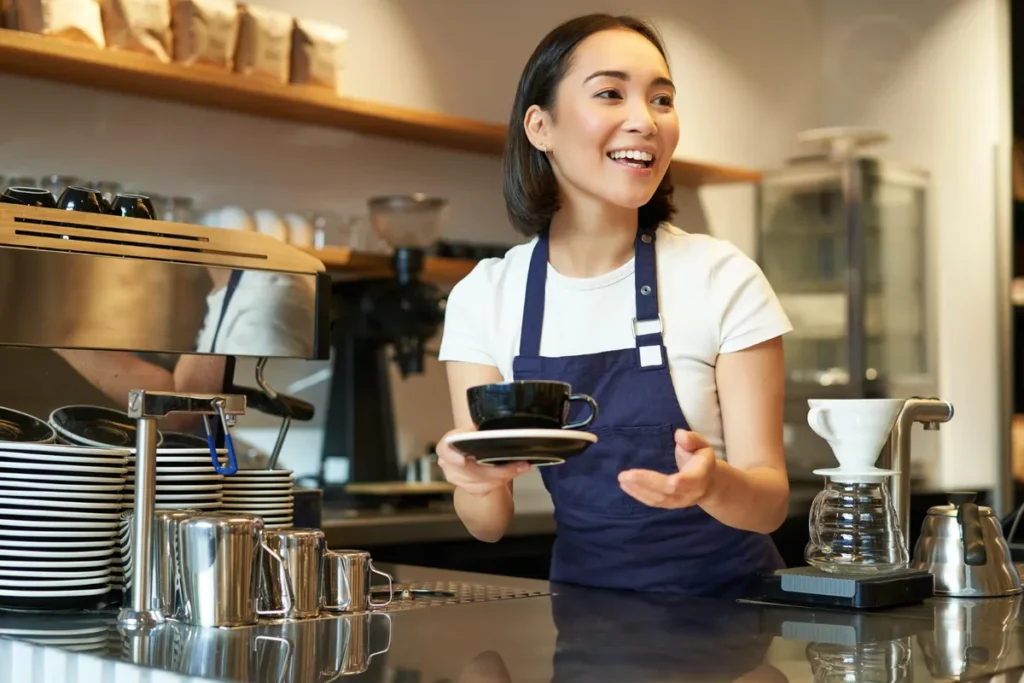 Barista (mulher asiática) em uma cafeteria gourmet, vestindo avental azul e camiseta branca, segurando uma xícara de café enquanto sorri. Equipamentos como um Hario V60 e máquina de espresso estão visíveis.